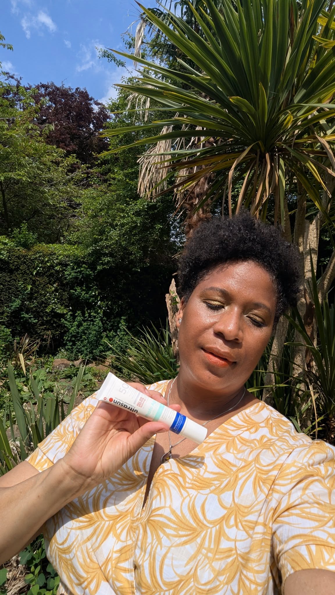 black woman with afro holding tube of Ultrasun Photo Age sun protection set againt palm tree back drop